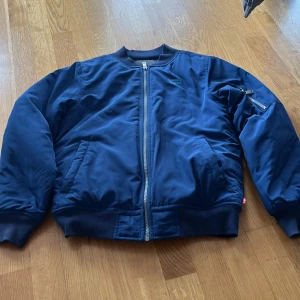 Blå bomberjacka från Levi's - Säljer en stilren blå bomberjacka från Levi's med dragkedja framtill och ribbade muddar. Jackan har fickor på sidorna och en extra ficka med dragkedja på ena ärmen. Perfekt för dig som vill ha en klassisk look. Stl L för barn, ca 11-13 år. 