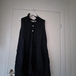 Svart ärmlös klänning med knappar - Svart ärmlös klänning med knappar framtill och lätt krage. Klänningen har en lös passform och är tillverkad i ett lätt, luftigt material. Perfekt för varma dagar eller att styla med accessoarer.