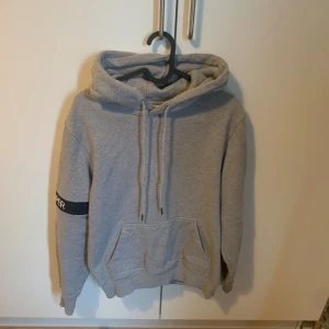 Grå hoodie av märket daily paper - Säljer en enkel grå hoodie med huva och känguruficka framtill. Tröjan har en svart detalj med text på vänster ärm och dragsko i huvan. Perfekt för en avslappnad stil.