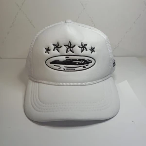 Vit keps med broderade stjärnor - Snygg vit keps med svart broderi av stjärnor framtill och på sidan. Kepsen har mesh-bakstycke och justerbar knäppning bak för perfekt passform. Framsidan har även ett broderat ovalt motiv.