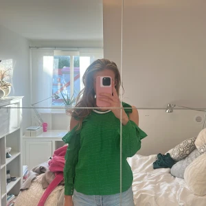 Grön offshoulder tröja med volang - Säljer en snygg grön tröja med offshoulder-design och volangdetaljer. Tröjan har lång ärm och är tillverkad i ett lätt, strukturerat material. Perfekt för dig som vill sticka ut med en färgglad och trendig look.