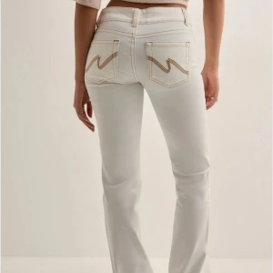 Vita bootcut jeansbyxor med kontrastsömmar. - Snygga vita jeansbyxor med bootcut-passform och hög midja. Byxorna har kontrastsömmar i beige, dubbla knappar i midjan och klassiska bakfickor med dekorativa sömmar. Perfekta för en trendig och fräsch look. Säljer dessa pga att dom är förstora, använt endast 2 gånger. OBS! Dom är lite missfärgade längst ner på byxorna pga att dom har släpats i marken när jag har gått!