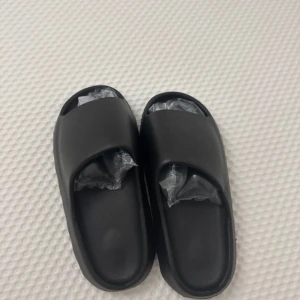 Svarta slides  - Svarta slides med bred rem över foten och öppen tå. Gjorda i syntetmaterial med en chunky, vågformad sula för extra komfort. Perfekta för sommaren eller som inneskor.