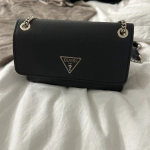 Svart axelväska från Guess - Snygg svart axelväska från Guess med silverfärgad kedja och logga framtill. Väskan har flera fack inuti, dragkedja och magnetlås. Perfekt storlek för det viktigaste och stilren design som passar till många outfits.