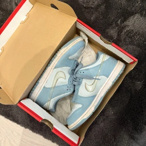 Nike Dunk Low i ljusblått och vitt - Säljer ett par Nike Dunk Low i snygg ljusblå och vit färg. Skorna har klassisk låg siluett, vit sula och ljusblå snörning. Perfekta för dig som gillar streetwear och vill ha en stilren sneaker i garderoben. 