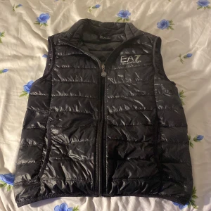 Svart dunväst från Emporio Armani EA7 - Snygg svart väst från Emporio Armani nypris 2500 EA7 med quiltad design och dragkedja framtill. Västen har hög krage och logga på bröstet. Perfekt för lager-på-lager och stilren look.