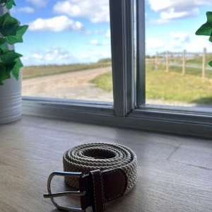 Beige flätat bälte med bruna skinn-detaljer - Snyggt beige flätat bälte med bruna skinn-detaljer vid spännet och silverfärgat metallspänne. Perfekt accessoar för att lyfta din outfit med en stilren touch. Passar både jeans och chinos.