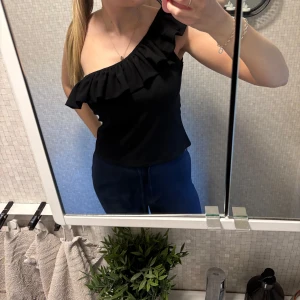 Svart oneshoulder topp med volang - Säljer en svart oneshoulder topp med volang, en väldigt snygg fest topp!🎉 storlek M, superfint skick!🩷