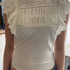Vit blus med spets och volang - Säljer en söt vit blus med spetsdetaljer och volanger på axlarna. Blusen har ett broderat blommönster upptill och är öppen i armarna. Inte så tajt i verkligheten som på bild