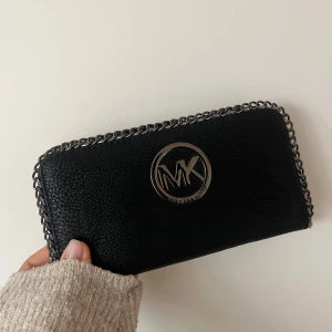 Svart plånbok från Michael Kors med kedjedetalj - Säljer en svart plånbok från Michael Kors. Plånboken är i skinnimitation och har dragkedja. Perfekt accessoar för att hålla ordning på kort och kontanter. Skriv privat för mer info eller bilder. Pris kan diskuteras vid snabb affär:)