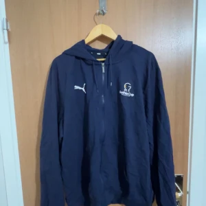 Marinblå hoodie från Puma x Gothia Cup - Säljer en marinblå hoodie från Puma med Gothia Cup-logga på bröstet. Jackan har huva med dragsko och dragkedja framtill. Perfekt för dig som gillar sportig stil och vill ha något bekvämt och snyggt.