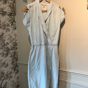 Jeansklänning Michael Kors - Säljer en jeansklänning med omlottdesign från Michael Kors. Klänningen har markerad midja med resår och dekorativa axelknappar. Perfekt för varma dagar och enkel att styla med sneakers eller sandaler.