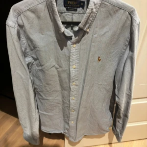 Ljusblå skjorta från Polo Ralph Lauren - Klassisk ljusblå skjorta från Polo Ralph Lauren med button-down krage och broderad logga på bröstet. Skjortan har långa ärmar och knäppning framtill. Perfekt för en stilren och tidlös look.