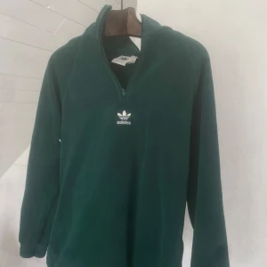 Mörkgrön fleece-tröja från Adidas - Mörkgrön fleece-tröja från Adidas med halv dragkedja och broderad logga på bröstet. Tröjan har lång ärm och hög krage, perfekt för kyligare dagar. Mjuk och skön känsla tack vare fleecematerialet.