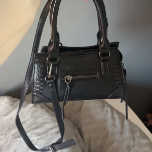 Svart handväska med axelrem - Säljer en svart handväska med både handtag och justerbar axelrem. Väskan har en dragkedja framtill och har två mindre fack i! 