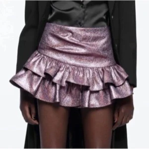 Glittrig Zara volangkjol i metallic lila - Supersnygg kortkjol i metallic lila med glittrig finish och dubbla volanger. Kjolen är från Zara och har en figurnära passform upptill och markerad midja. Perfekt för dig som vill sticka ut med en trendig och cool look.