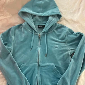 Ljusblå velourbyxor från Juicy Couture - Säljer ett par ljusblå velourbyxor från Juicy Couture med raka ben och resår i midjan. Byxorna har fickor fram och bak samt en liten broderad logga framtill. Tröjan är storlek M och byxorna storlek L