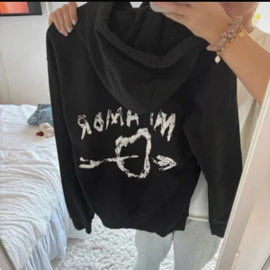 Svart hoodie med tryck på ryggen - Säljer en svart hoodie med vit text och pilhjärta-tryck på ryggen. Klassisk modell med huva och känguruficka framtill. Perfekt för en avslappnad stil.