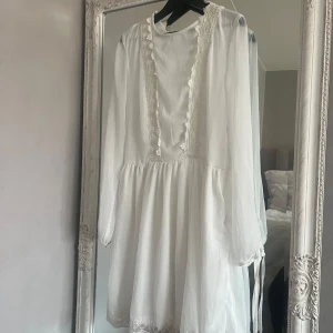 Vit spetsklänning med snörning - Romantisk vit klänning med spetsdetaljer längs halsringningen och nederkanten. Långärmad modell med v-ringning och dekorativ snörning i ryggen. Perfekt för sommarens tillfällen eller studentfirande. Storlek 40 men sitter mera som en 38/36
