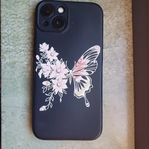 Svart mobilskal med fjäril och blommor IPhone 13 - Säljer ett svart mobilskal med en stor dekorativ fjäril och rosa blommor på baksidan. Skalet har en matt finish och motivet är detaljerat med inslag av vitt, rosa och gult. Perfekt för dig som vill ha ett unikt och snyggt mobilskal.