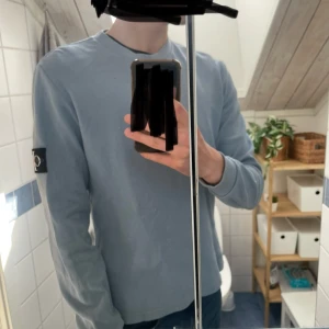 Ljusblå långärmad tröja från Calvin Klein Jeans - Säljer en ljusblå långärmad tröja från Calvin Klein Jeans med stort CK-märke på ärmen. Tröjan har rund hals och är tillverkad i ett strukturerat bomullstyg som ger en stilren och avslappnad look. Ny pris 600 mitt pris 299