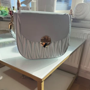 Ljusgrå quiltad axelväska med gulddetaljer - Säljer en ljusgrå axelväska med quiltad design och snyggt guldfärgat spänne framtill. Väskan har en kedja som axelrem och är perfekt för att lyfta vilken outfit som helst.