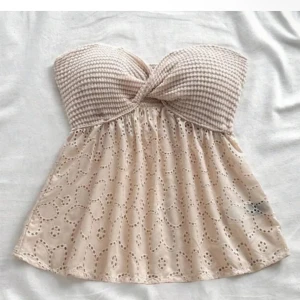 Beige axelbandslös topp med broderade detaljer - Söt beige axelbandslös topp med twistad byst i ribbat material och broderad, lätt utsvängd nederdel med blommönster. Perfekt för varma dagar och en avslappnad stil.