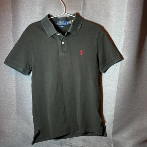 Svart piké t-shirt från Polo Ralph Lauren - Svart Piké från Polo Ralph Lauren. Piken är lite solblek så den har tappat lite färg. Storlek 14-16 år. Skick 8/10. Stryker innan vi fraktar.