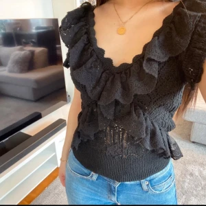 Svart volangtopp med spetsdetaljer - Säljer en svart topp med volanger och spetsdetaljer. Toppen har djup v-ringning, är ärmlös och har en åtsittande midja. Perfekt för dig som vill ha en feminin och trendig look.