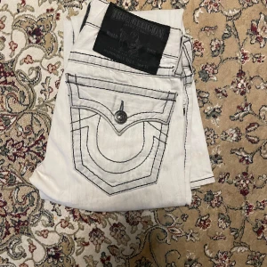Vita jeansbyxor från True Religion - Säljer ett par vita jeansbyxor från True Religion med svarta kontrastsömmar och klassiska bakfickor med lock och knapp. Byxorna har raka ben och tydliga detaljer som logga på bakfickan och patch vid linningen. Har små gulaktiga fläckar längst ner men märks inte när man har de. Har även lite healbite