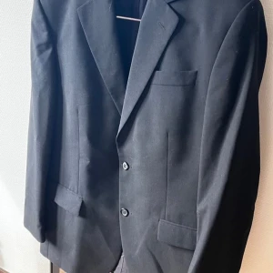 Svart kavaj från Wilhelm - Klassisk svart kavaj från Wilhelm med två knappar framtill och tre fickor. Tillverkad i en blandning av ull och polyester, vilket ger en stilren och bekväm känsla. Perfekt för formella/casual tillfällen. Kavajen har två knappar fram och fyra knappar på varje ärmslut. Den är vintage så kavajen är oversize samt har axelvaddar.  Använt ett fåtal gånger, köptes i en second hand butik i Köpenhamn 