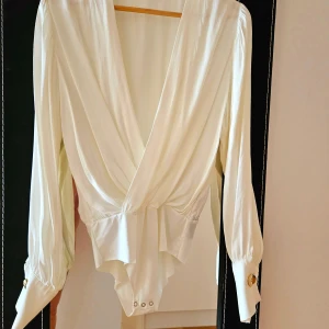Vit omlott bodysuit med långa ärmar - Säljer en elegant vit bodysuit med djup omlott-ringning och långa ärmar. Bodyn har breda manschetter med guldknappar och knäppning i grenen. Perfekt för en stilren look.