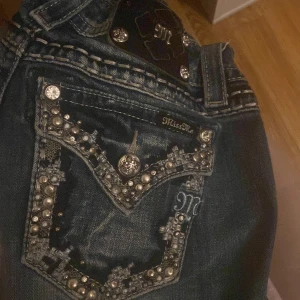 Miss Me jeans med pärldetaljer - Säljer ett par blå Miss Me jeans med snygga pärlor och glittriga detaljer på bakfickorna. Jeansen har låg midja och klassisk femficksmodell med dekorativa knappar och broderier. Perfekta för dig som vill sticka ut lite extra!