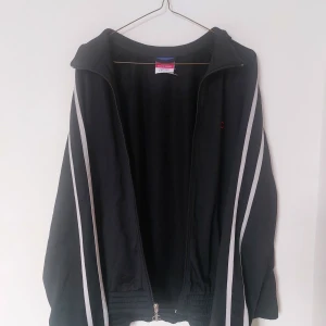 Svart zip-up jacka från Champion - Säljer en svart träningsjacka från Champion med vita ränder längs ärmarna och dragkedja framtill. Det är en Champion-logga på dragkedjan, samt en röd logga på börstet. Köpt secondhand, fint skick. Pris kan diskuteras!
