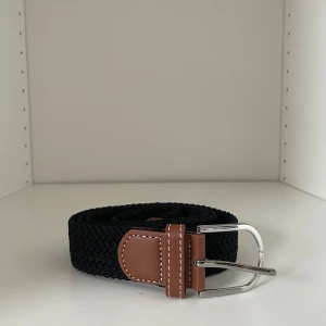 Svart flätat bälte med bruna skinn-detaljer - Snyggt svart flätat bälte med bruna skinn-detaljer och silverspänne. Perfekt accessoar för att lyfta din outfit. Bältet har klassisk design med kontrastsömmar på skinnet och passar till många olika stilar. Bältet är 110cm