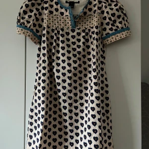 Mönstrad kortklänning med hjärtan och blå detaljer - Supersöt kortklänning med blå hjärtmönster på beige botten. Klänningen har puffärmar, blåprickiga kantband och dekorativa partier med småmönster. Perfekt för dig som gillar unika detaljer och lekfull stil.