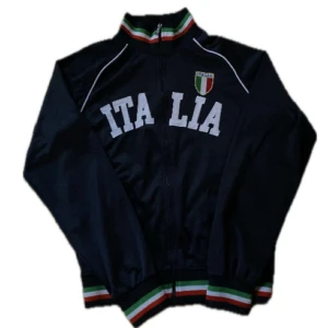 Chief keef zip - Säljer en svart track jacket med vita bokstäver 'ITALIA' och broderad italiensk sköld på bröstet. Jackan har dragkedja, ribbade muddar och krage med Italiens färger. Perfekt för dig som gillar sportig stil.  Storleken är xl men skulle säga att den passar som medium