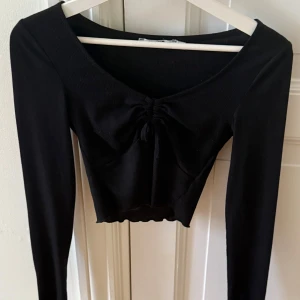 Svart croppad topp från NA-KD - Den här toppen är ribbad och har långa ärmar med en babylock-design, en dragsnöresdetalj på bröstet, en söm i babylock-stil på nederfållen, en croppad längd och ett mjukt material. Den är V-ringad och perfekt för en utekväll! 