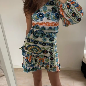 Mönstrad one shoulder klänning med volang - Säljer en färgglad kortklänning med blommigt och geometriskt mönster i blått, grönt, orange och vitt. Klänningen har en one shoulder-design med lång ärm och volangdetaljer längs nederkanten och ärmslutet. Perfekt för dig som gillar unika plagg!