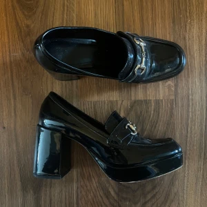 Svarta platåloafers med klack - Säljer ett par svarta loafers med hög klack och platåsula. Skorna har en glansig finish och dekorativa metallspännen framtill. Perfekta för dig som vill ha en klassisk men edgy look.