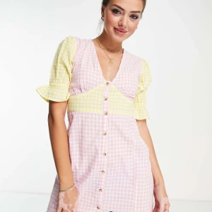 Rutig klänning med puffärm och knappar - Söt klänning med rosa och vit rutig kjol och liv, samt gula och vita rutiga puffärmar. Djup v-ringning, knappar framtill och kort ärm med volangkant. Perfekt för vår och sommar.