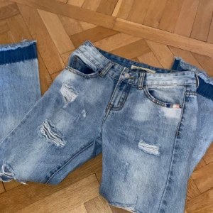 Ljusblå slitna jeans med råa kanter - Säljer ett par ljusblå jeans med slitningar och hål på framsidan. Byxbenen har en mörkare blå kant nertill och råa, fransiga avslut. Klassisk femficksmodell med normal passform och hög midja.