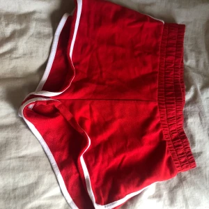 Röda sportiga shorts med vit kant - Snygga röda shorts med elastisk midja och vita kanter längs benen och sidorna. Perfekta för träning eller en avslappnad stil. Mjukt material och klassisk sportig look.
