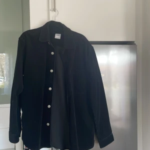 Svart manchester overshirt från Zara - Säljer en svart overshirt i manchester från Zara. Skjortan har långa ärmar, klassisk krage och silverfärgade knappar framtill. Perfekt att bära som lager över en t-shirt eller tröja.