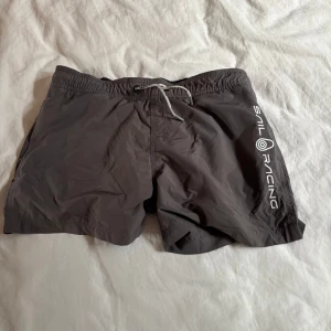 Svarta Sail Racing badshorts med snörning - Svarta badshorts från Sail Racing med vit logga längs ena benet. Shortsen har elastisk midja med snörning och meshfoder på insidan. Perfekta för stranden eller poolen, klassisk och sportig look.
