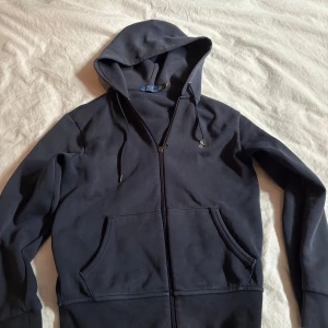 Mörkblå hoodie från Polo Ralph Lauren - Mörkblå hoodie från Polo Ralph Lauren i klassisk modell med dragkedja framtill, huva med snörning och två stora fickor. Tillverkad i mjuk bomull, perfekt för chill dagar. Snygg och enkel att matcha med jeans eller joggers. Den har en fläck på framsidan men de är inget som syns jätte mycket.