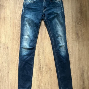 Replay Jeans - Snygga jeans från Replay i mörkblå tvätt med slitningar och ljusa partier. Klassisk femficksmodell med broderade bakfickor och Replay-logga på fickan. Jeansen är slim Fit, och är i storleken W25 L30. Jeansen är endast använda ett fåtal gånger.