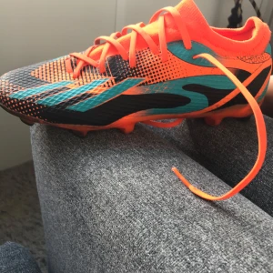 Färgstarka Messi fotbollsskor -  Kan också byta mott nått Säljer ett par Adidas Messi fotbollsskor i orange, svart och turkos med coola grafiska mönster. Skorna har snörning, platt sula med dobbar och är tillverkade i syntetmaterial för bästa grepp på planen. Perfekta för dig som vill sticka ut på fotbollen!
