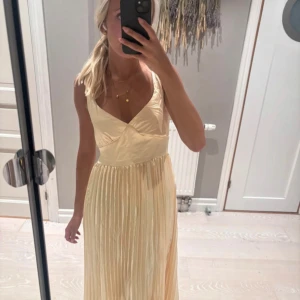 Champagnefärgad långklänning med öppen rygg - Superfin långklänning i champagnefärg med plisserad kjol och omlottliv. Klänningen har smala axelband och en snygg öppen rygg med korsade band. Materialet är glansigt och känns lyxigt, perfekt för speciella tillfällen som till exempel bal eller till bröllop. Klänning är aldrig använd. 