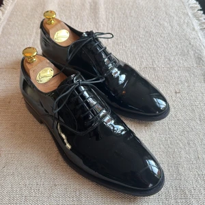 Svarta lackskor från Oscar Jacobson - Klassiska svarta lackskor från Oscar Jacobson med snörning och rund tå. Skorna är tillverkade i blankt skinn och har en låg klack samt lädersula. Perfekta för dig som vill ha en stilren och dressad look.
